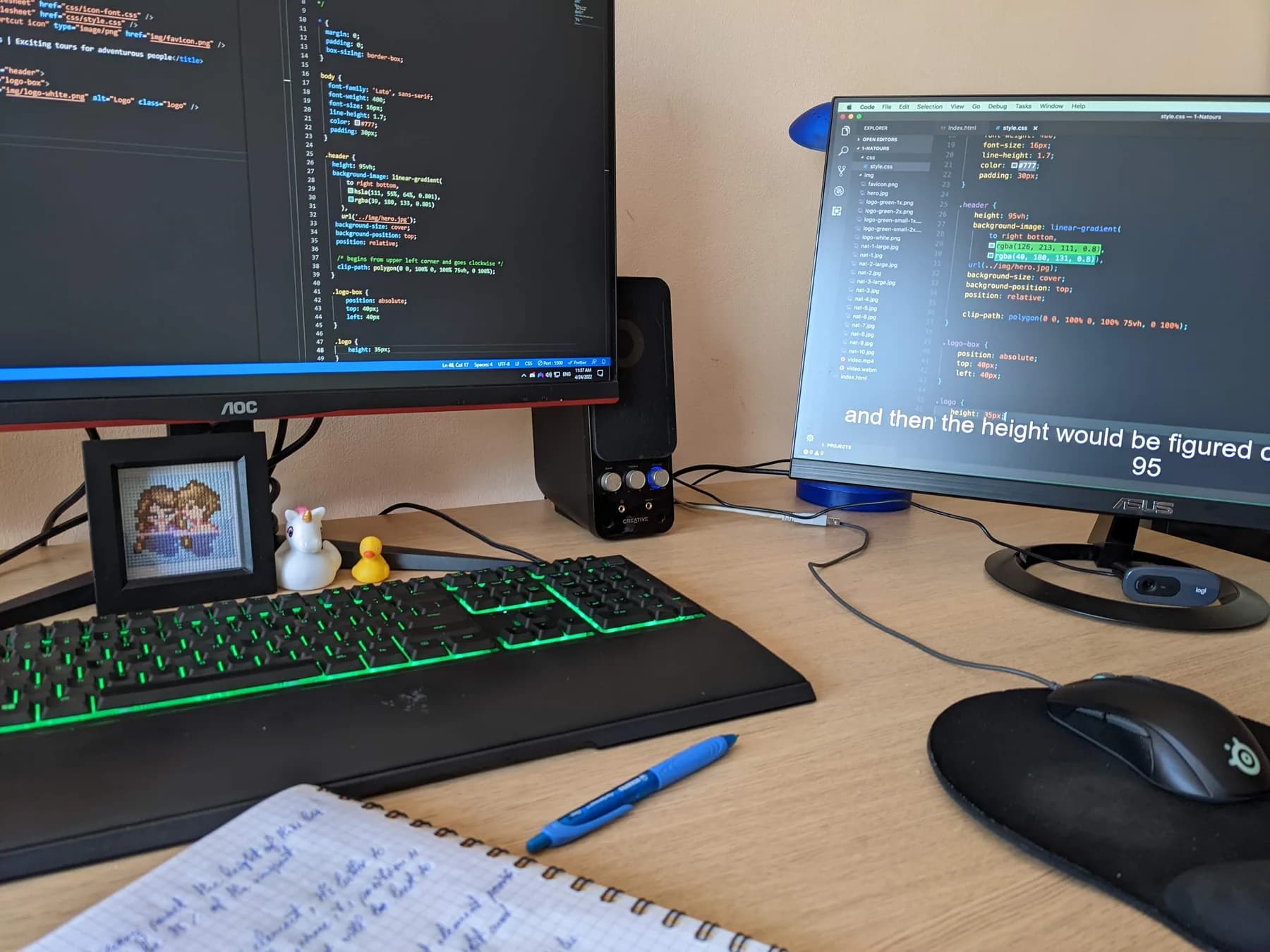 Coding desk with two monitors and Kami’s framed cross-stitch beside the keyboard